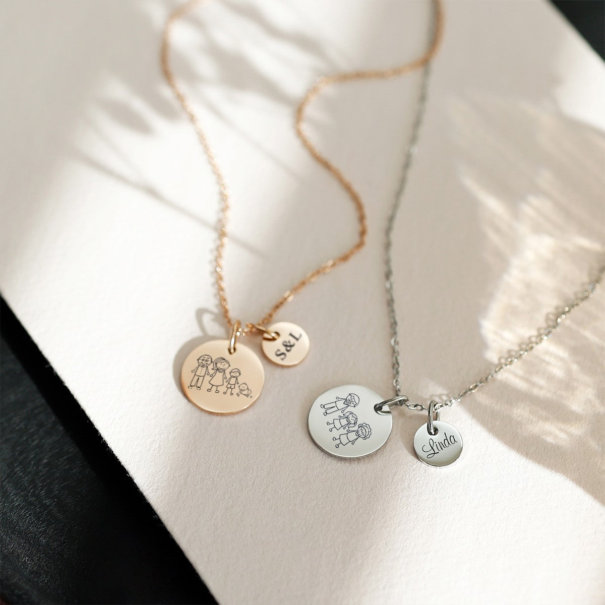 Personalisierte Familienkette in Roségold mit vierköpfiger Familie und Initialen-Plättchen S&L neben einer Familienkette in Silber mit drei Figuren und Namensplättchen Linda