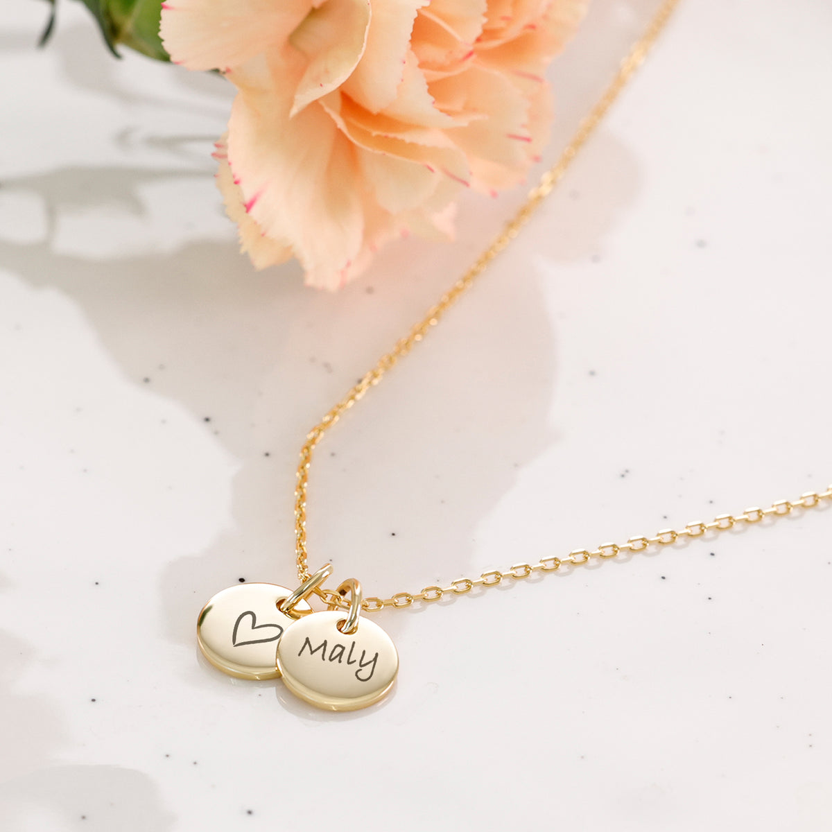 Goldkette mit zwei Plättchen, Herzmotiv und Gravur 'Maly', neben einer hellen Nelke